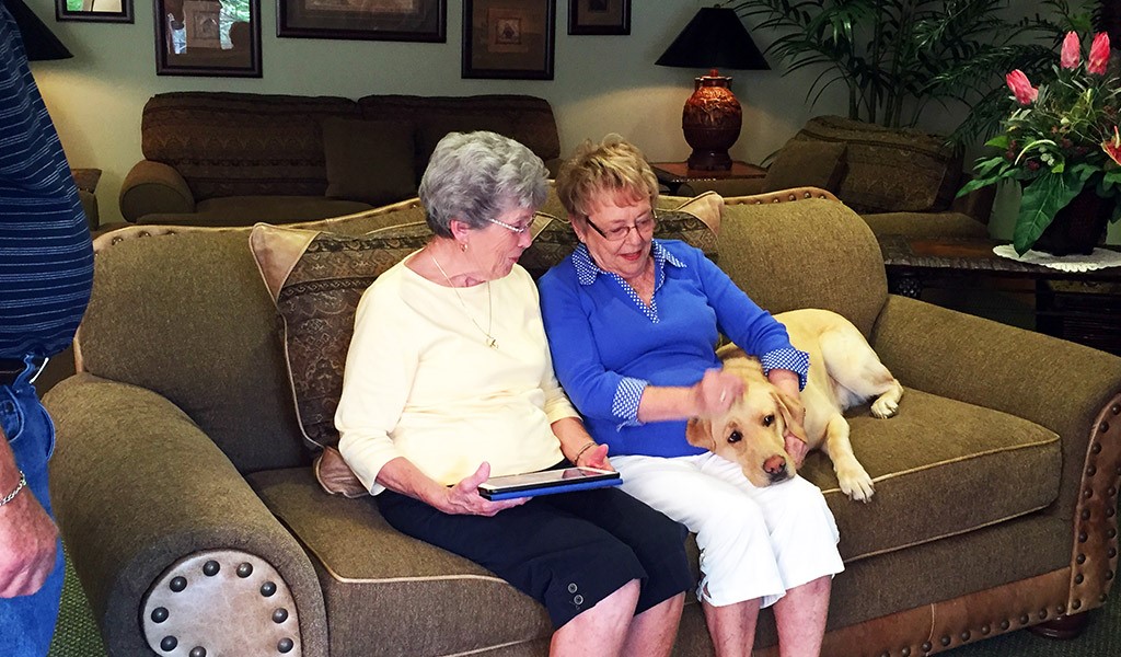 Two elderly women are seated on a couch, happily chatting, while a dog lies peacefully in their midst.